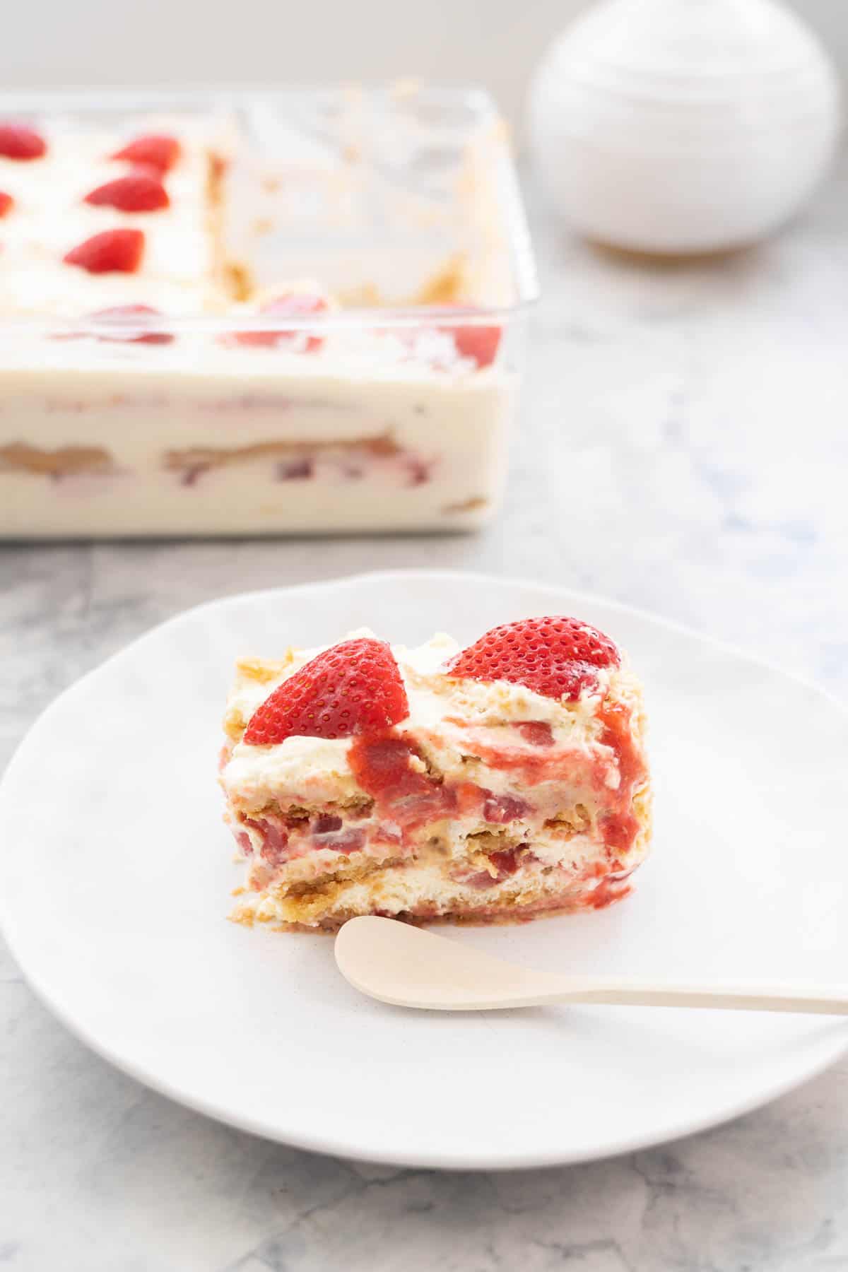A piece of a strawberry shortcake ice box cake on a plate with a white bamboo spoon in front of a glass container of more strawberry shortcake. 