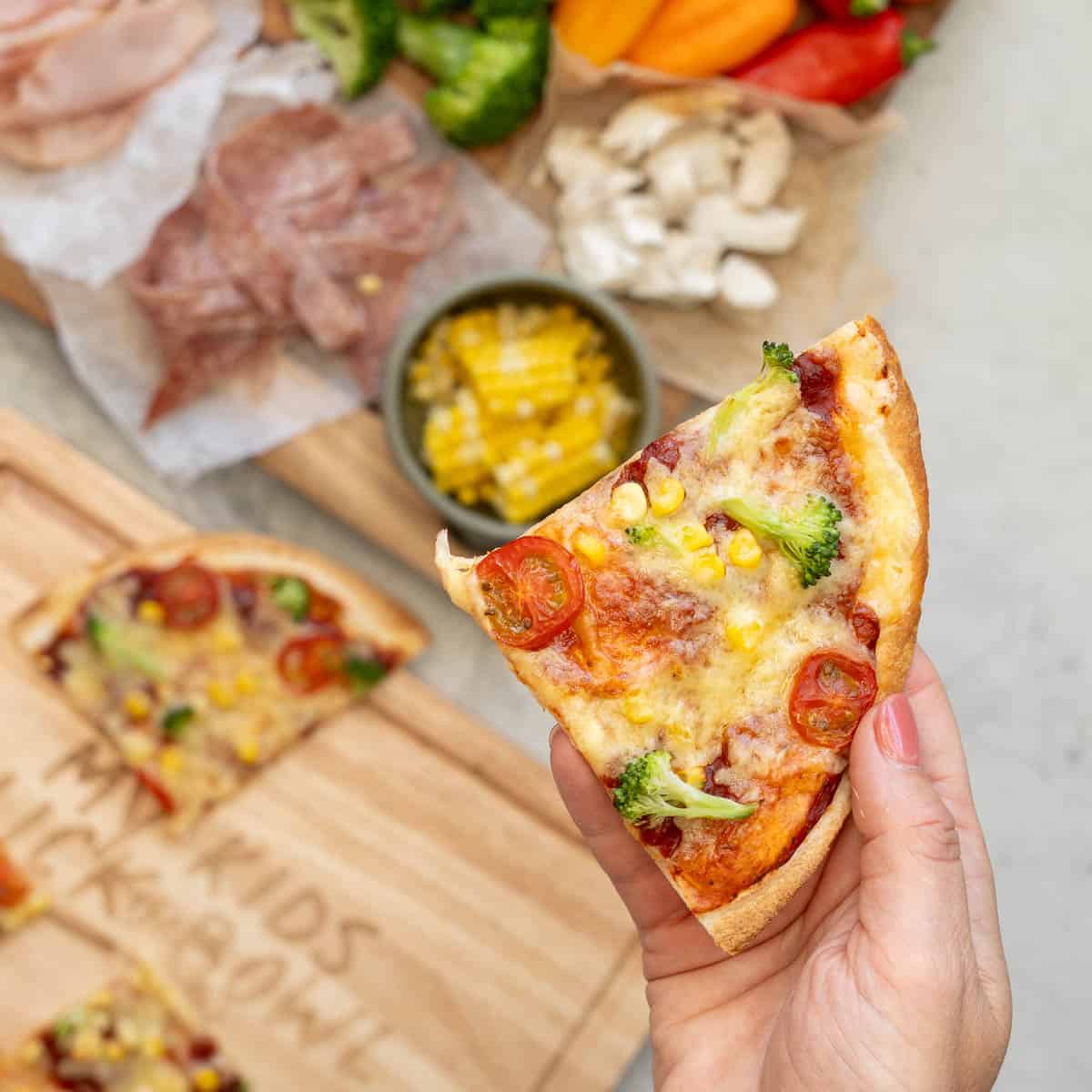 A hand holding up a slice of tortilla pizza above wooden serving boards with extra slices of pizza and pizza toppings 