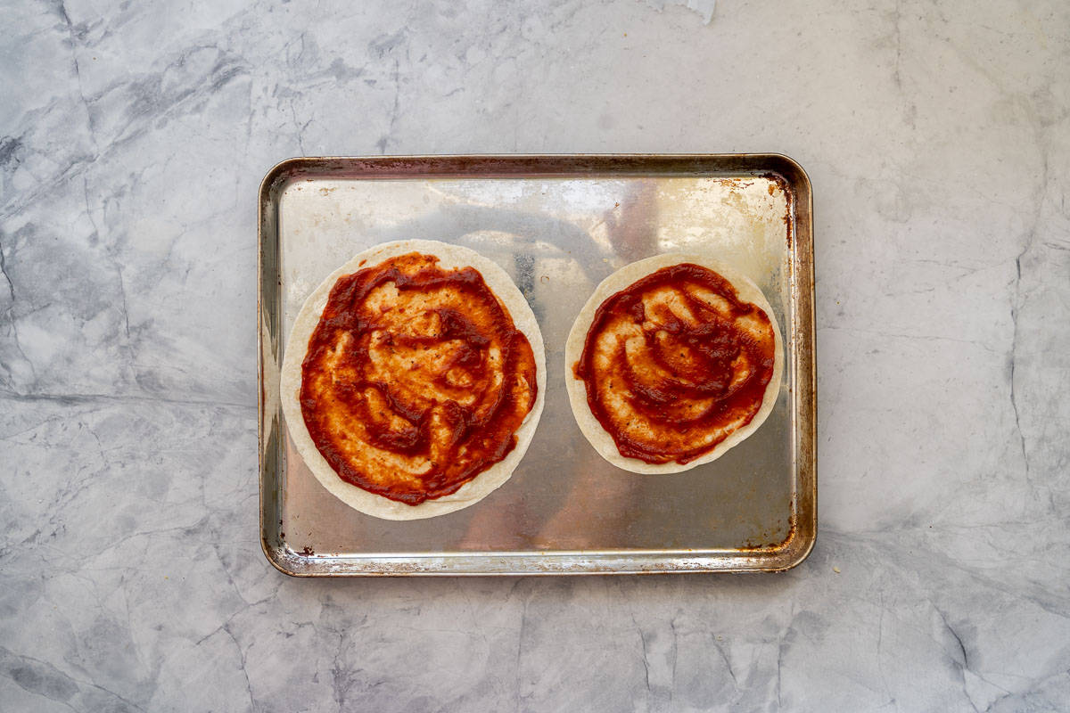 Two tortillas spread with pizza sauce on a baking tray resting on the bench 