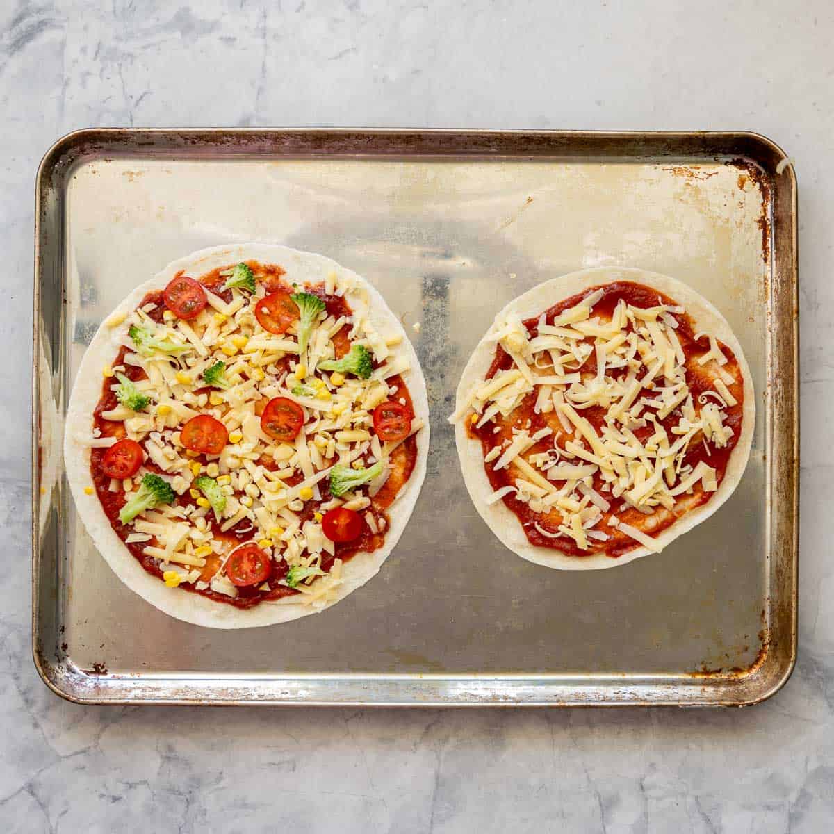 Two tortillas on a baking tray,  spread with pizza sauce and toppings and cheese