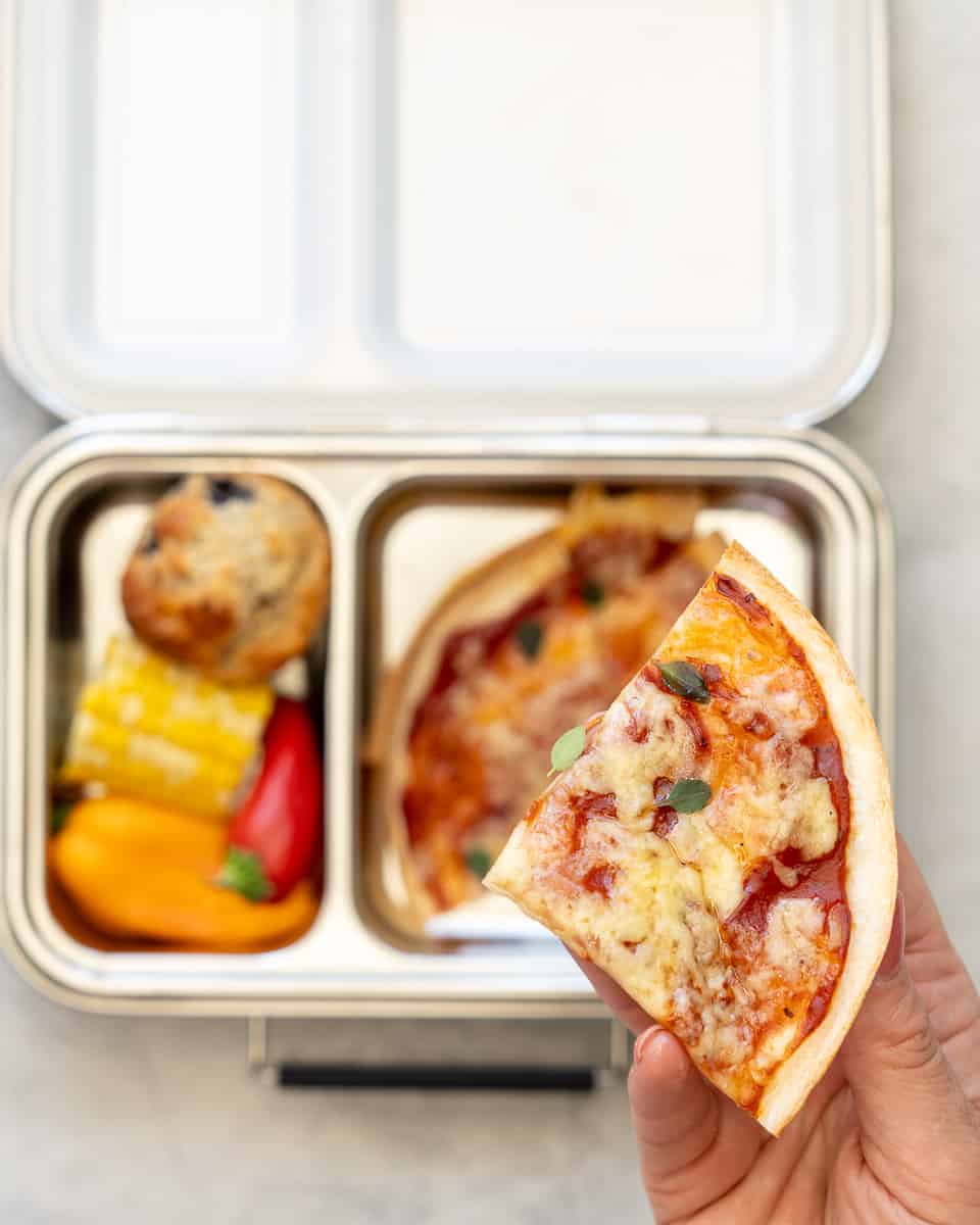 A hand holding up a slice of pizza above a tin lunch box below which is filled with more slices of the pizza , vegetables and a muffin.