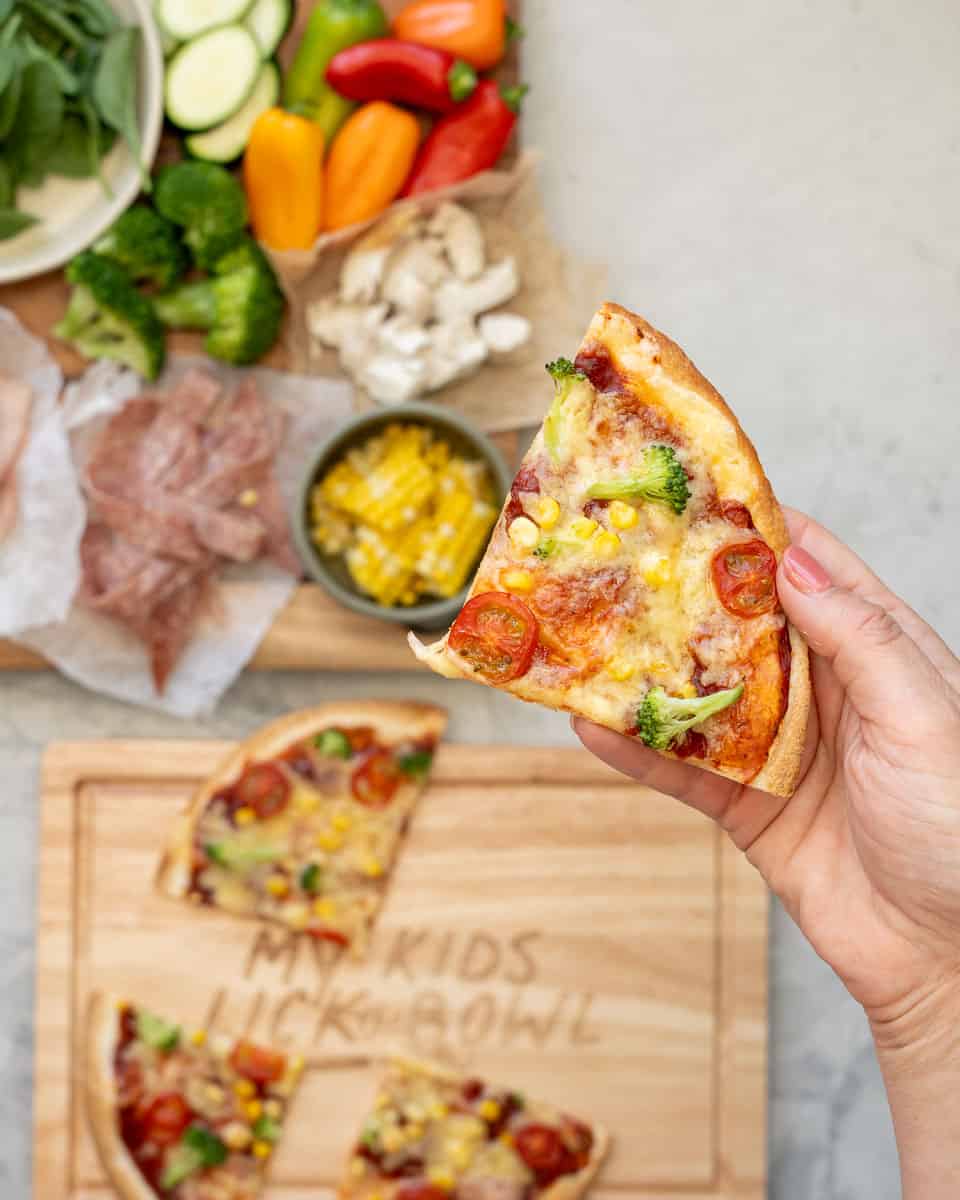 a hand holding up a slice of tortilla pizza above the rest of the pizza below which is on a serving board next to sliced vegetable toppings and a jar of sauce