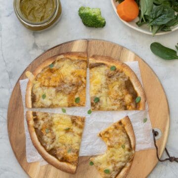 A wooden chopping board with a baked and sliced pizza on top of it, sitting next to a jar of vegetable pizza sauce and a bowl of fresh vegetables.