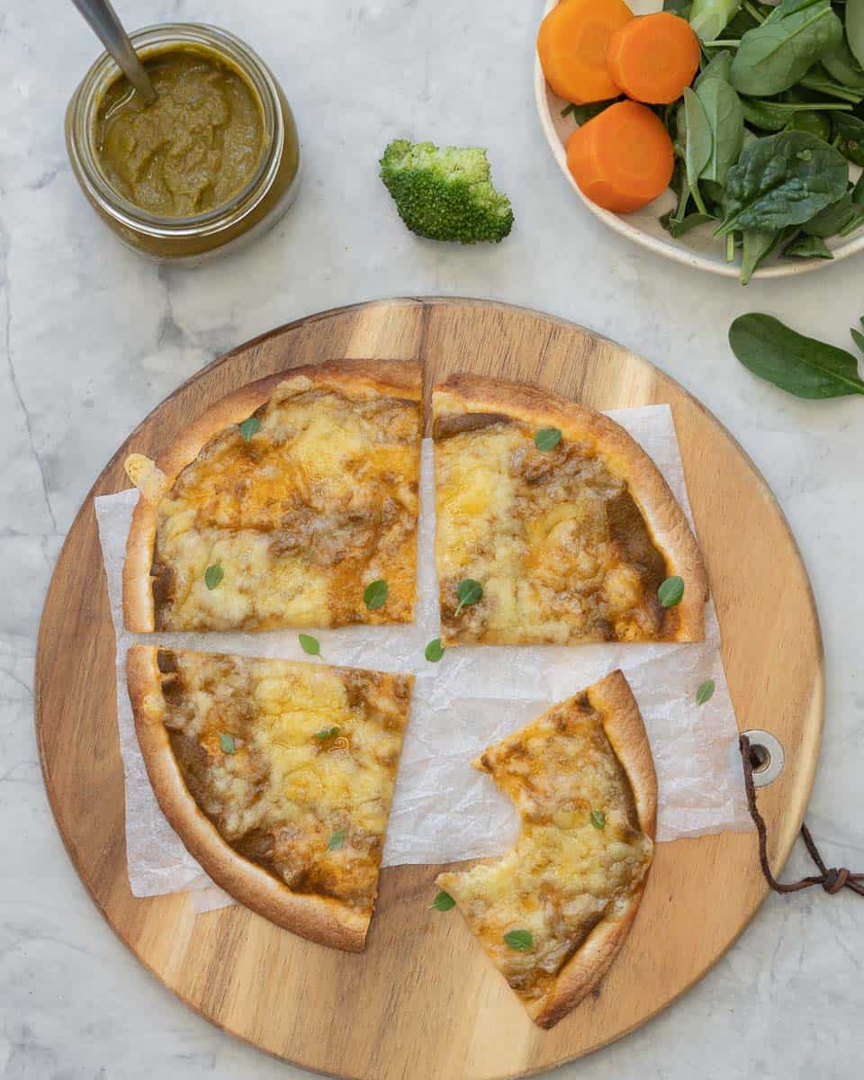 A wooden chopping board with a baked and sliced pizza on top of it, sitting next to a jar of vegetable pizza sauce and a bowl of fresh vegetables. 