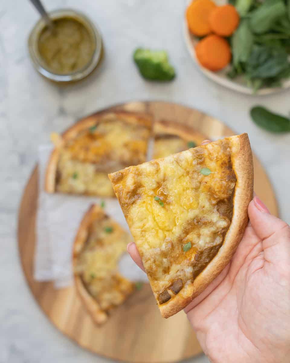 a hand holding up a slice of tortilla pizza above the rest of the pizza below which is on a serving board next to sliced vegetable toppings and a jar of sauce