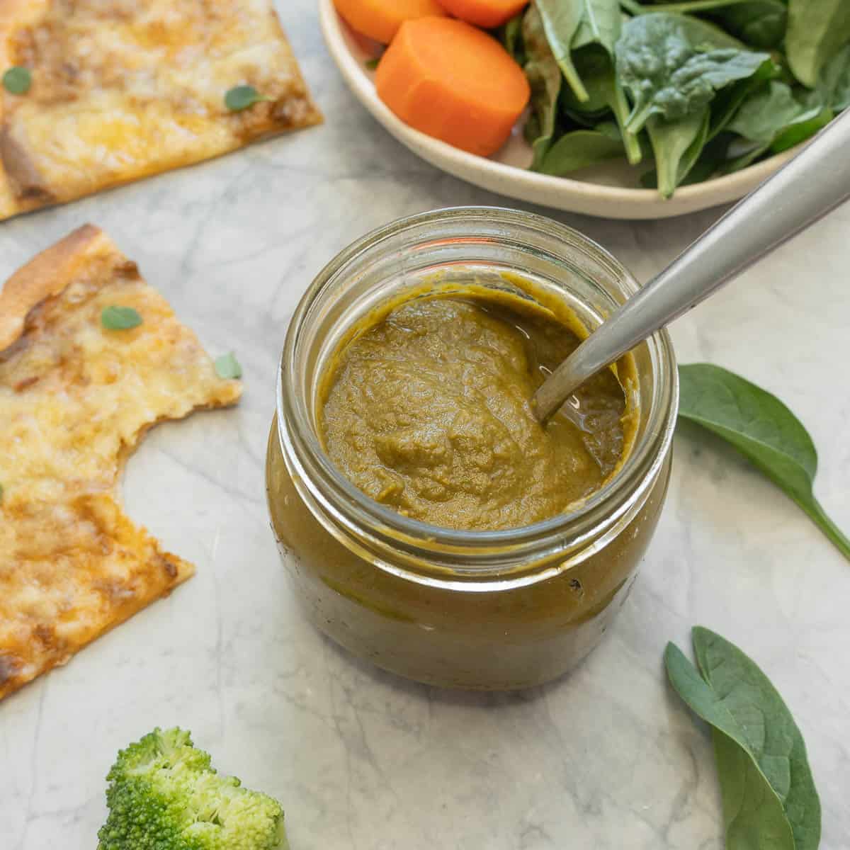 A small jar of vegetable pizza sauce sitting amongst a bowl of fresh vegetables, scattered spinach leaves and a pizza slices, one with a bite taken out of it. 