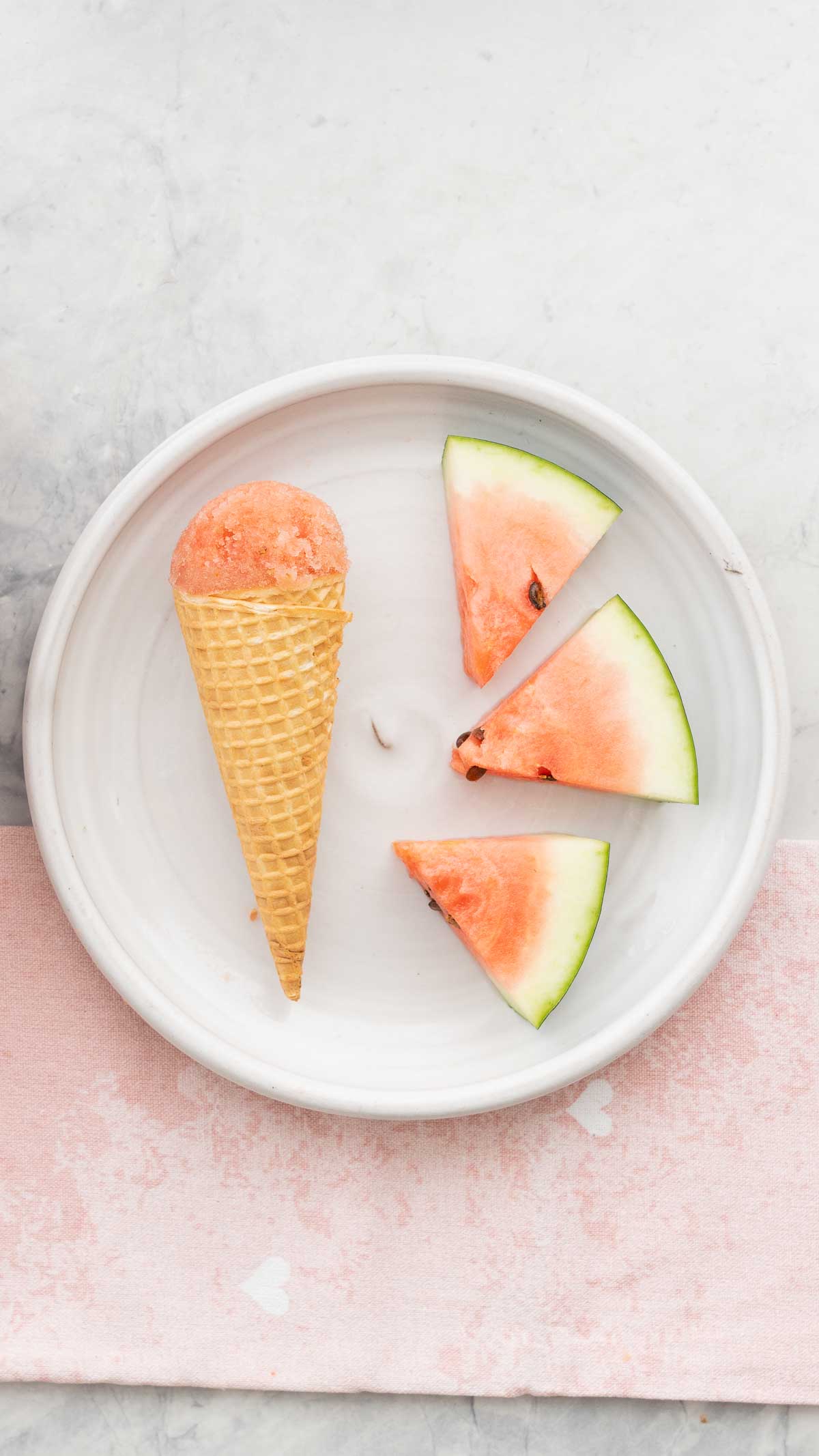 A waffle cone topped with a scoop of watermelon sorbet resting on a white plate with 3 watermelon slices. 