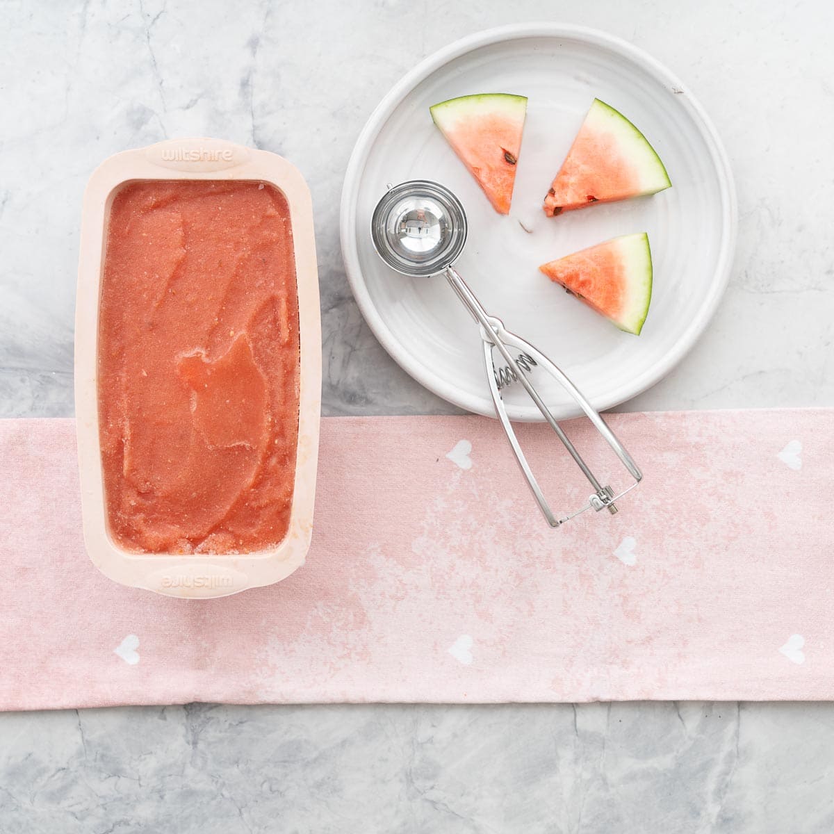 A loaf tin of watermelon sorbet next to a white plate with watermelon slices and an ice cream scoop. 