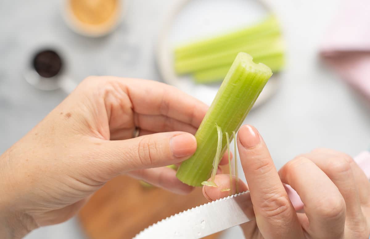 A pair of hands destringing a celery stick with a knife. 