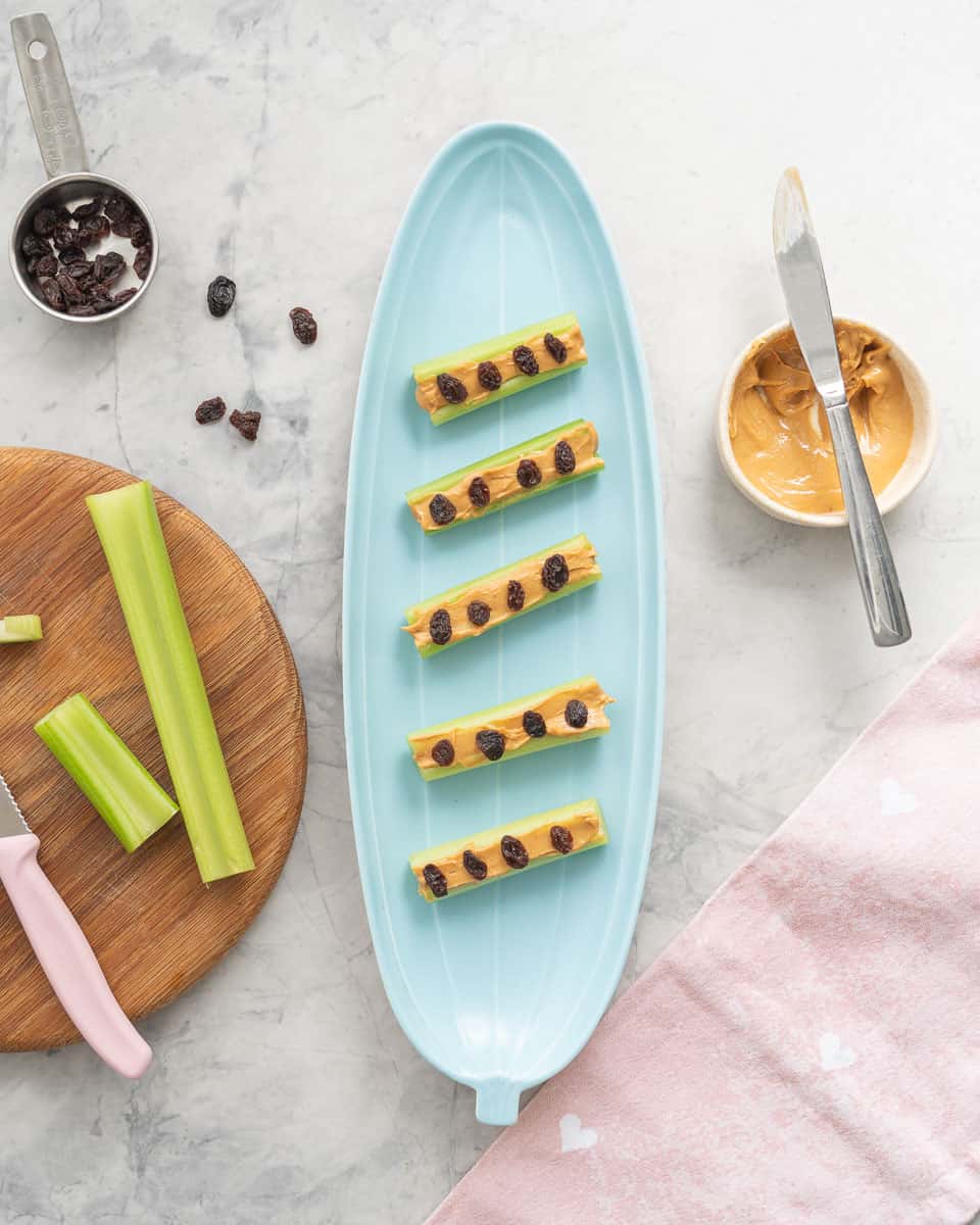 A plate full of 'ants on a log' served on an oval shaped platter which is next to a small ramekin of peanut butter and a wooden chopping board with extra sliced celery sticks