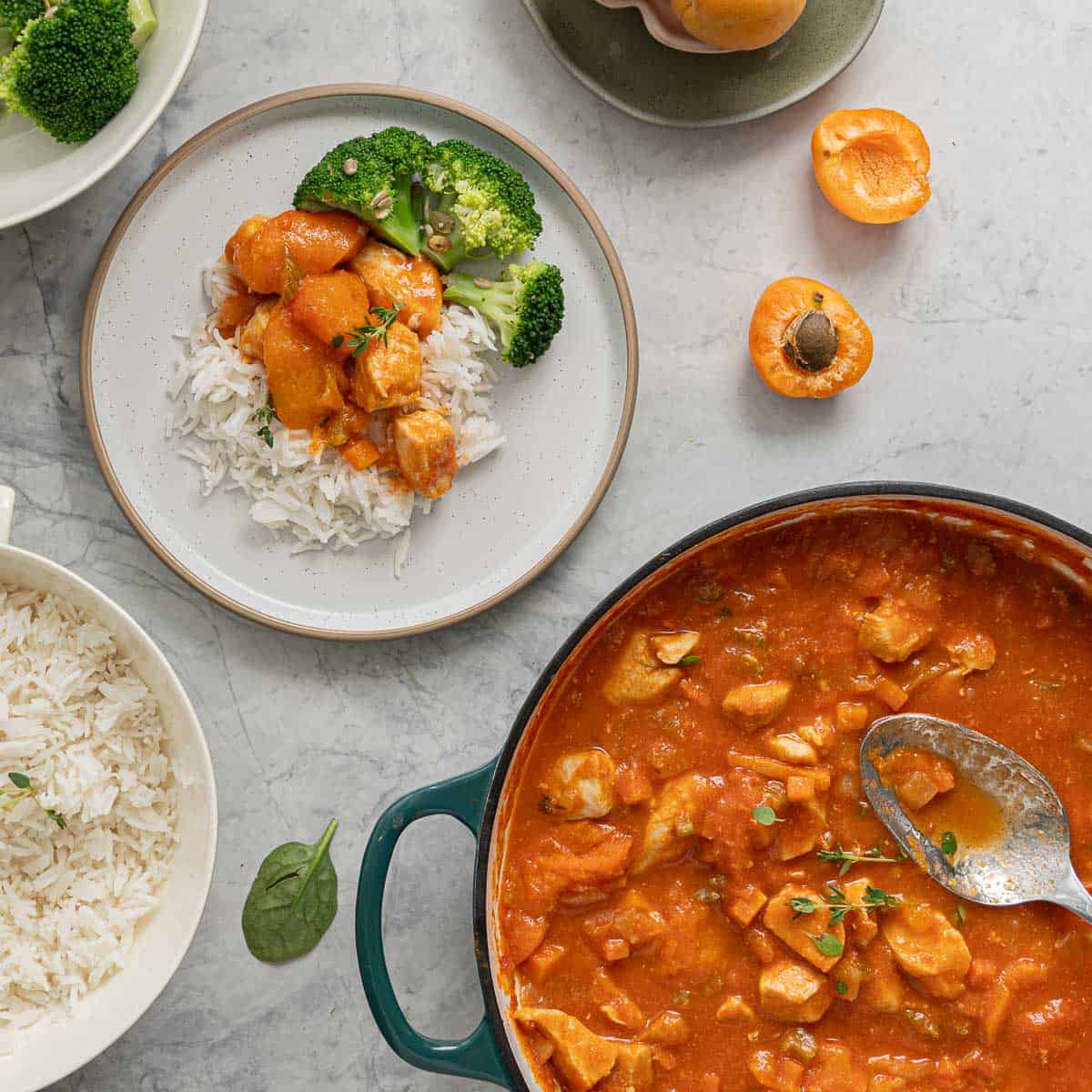 A pan full of apricot chicken, a bowl of rice and a bowl of broccoli sitting on the bench alongside fresh halved apricots and a serving on a plate. 