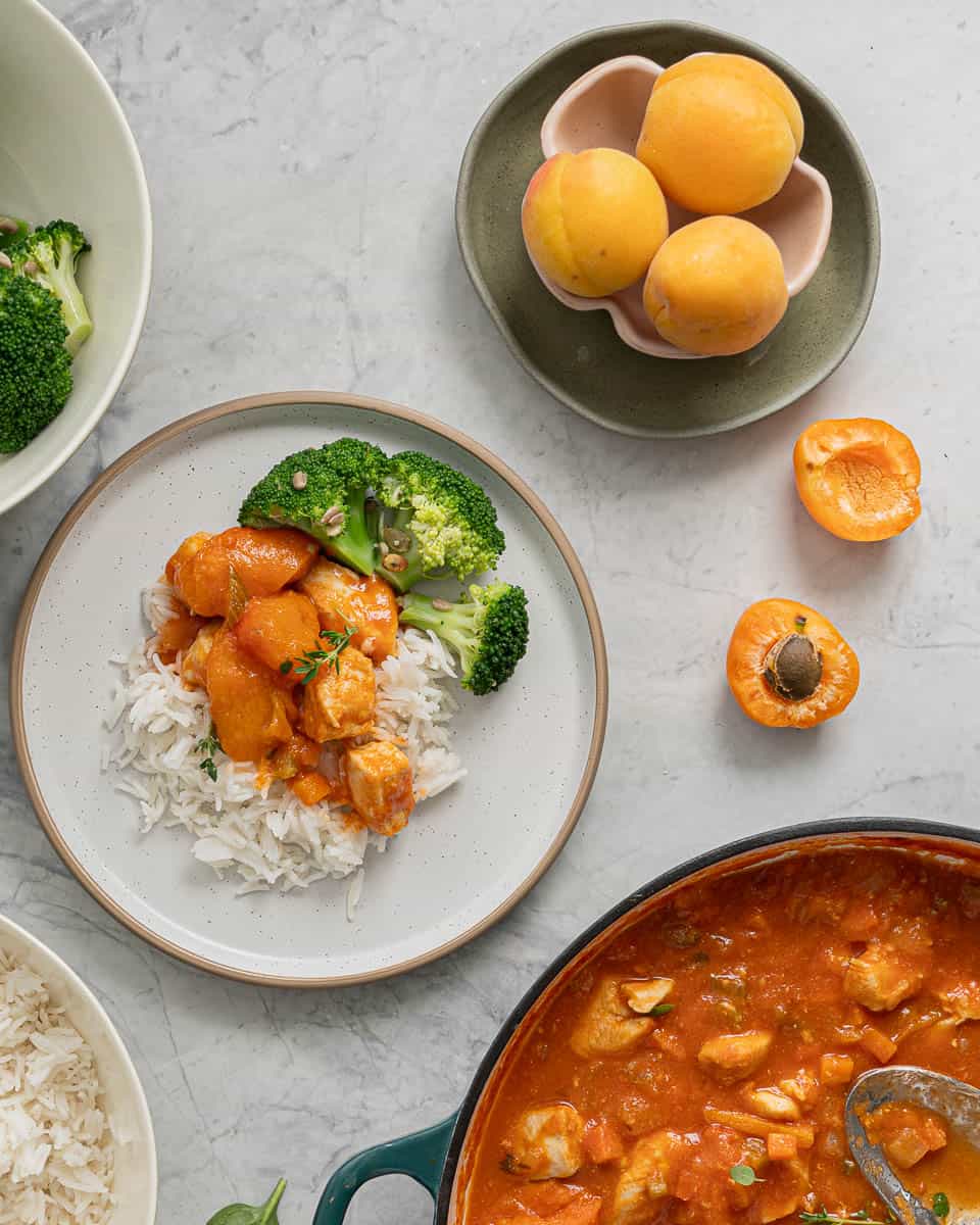 A serving of apricot chicken on a bed of rice and a side of broccoli which is sitting on the bench next to a bowl of fresh apricots and one which is sliced in half and resting on the bench