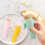 A banana popsicle in a aqua blue silicone squeeze tube being held above a plate of ice and more popsicles.