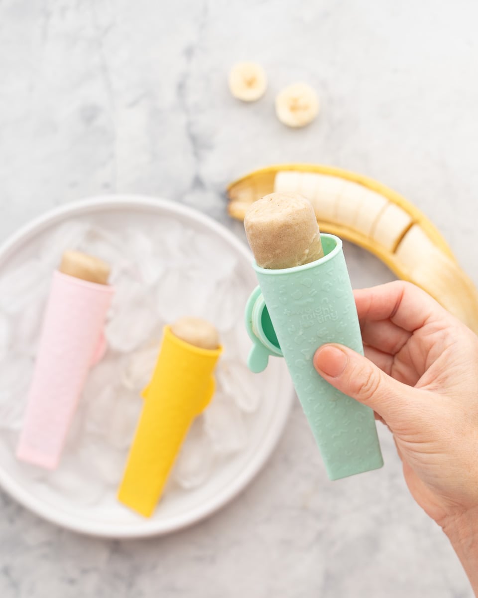 A hand holding a banana popsicle in a silicon mould above two popsicles below which is next to a sliced banana in its skin on the bench 