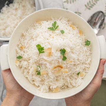A large bowl of Basmati rice, sprinkled with parsley and a scattering of toasted almonds, held above the bench by a pair of hands