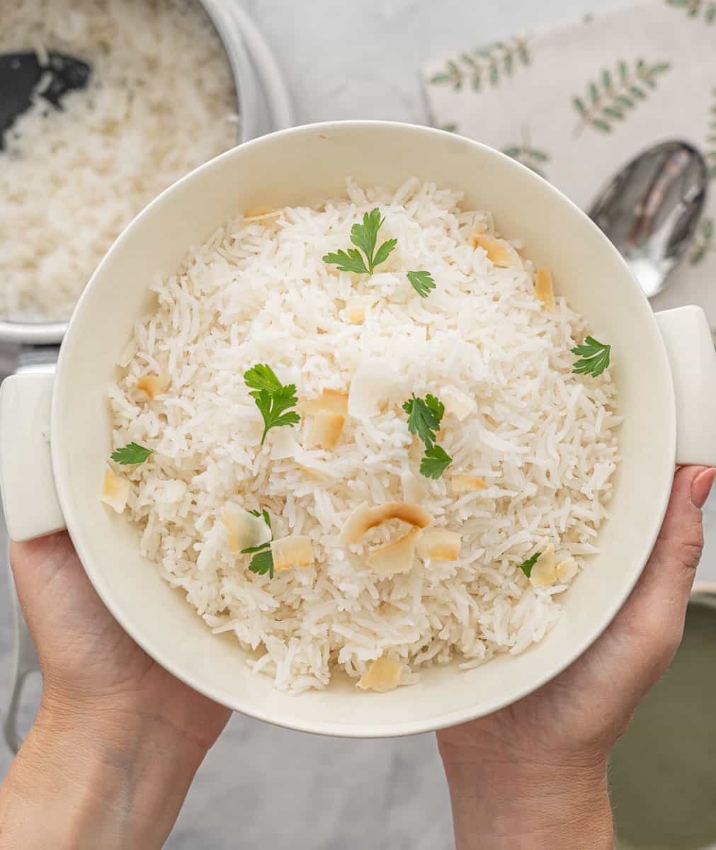 A large bowl of Basmati rice, sprinkled with parsley and a scattering of toasted almonds, held above the bench by a pair of hands 