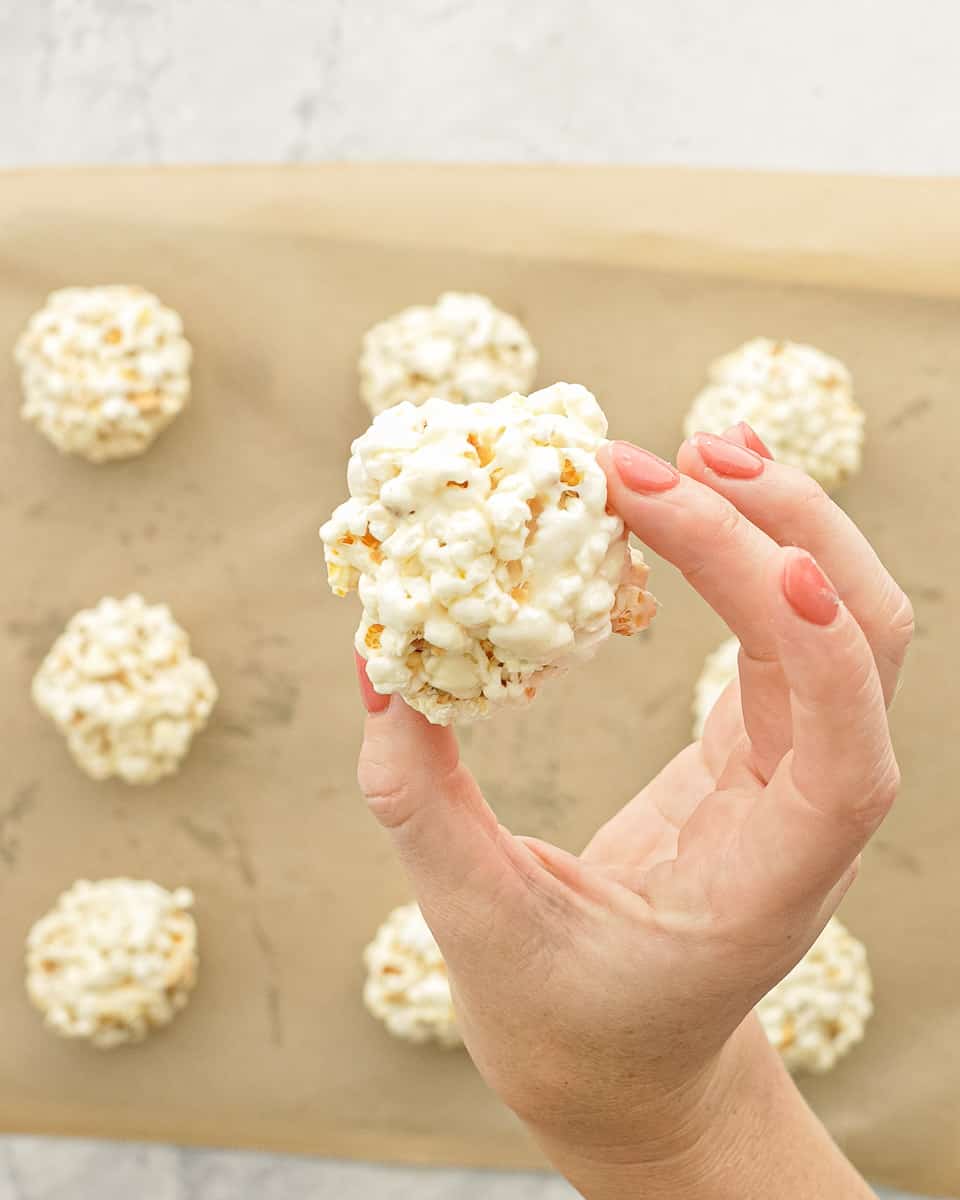 A hand holding a popcorn ball up to the camera above other popcorn balls resting on a sheet of parchment paper.