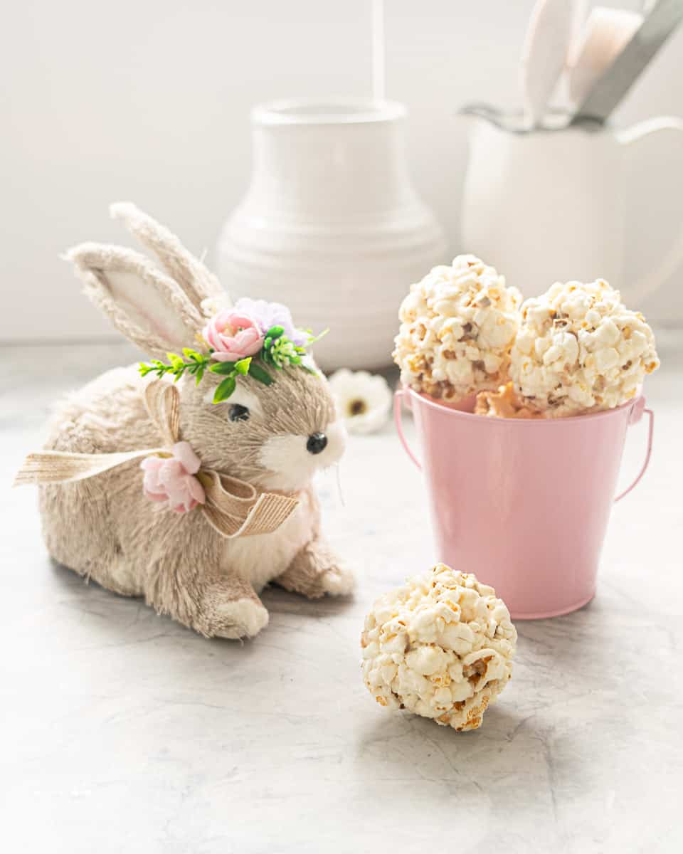 A tennis ball sized ball of popcorn on a bench in front of a decorative easter bunny and small pink bucket of more popcorn balls