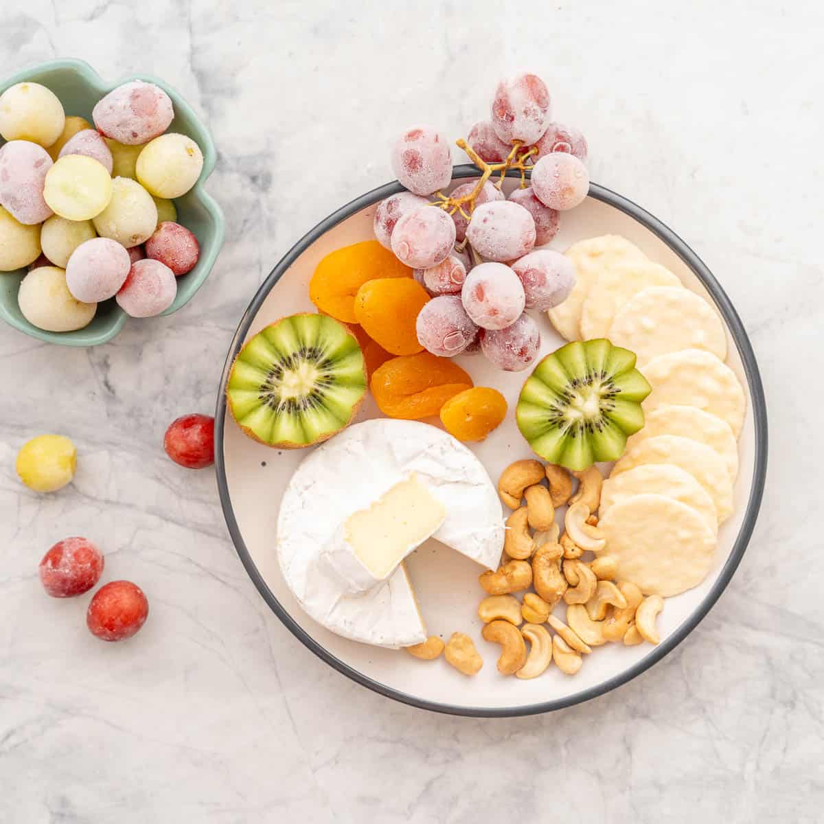A cheese, cracker and fruit platter with a bunch of frozen red grapes resting on the bench next to another bowl of frozen red and green graes. 