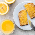 A jar of lemon curd and a piece of toast on a blue plate spread with lemon curd