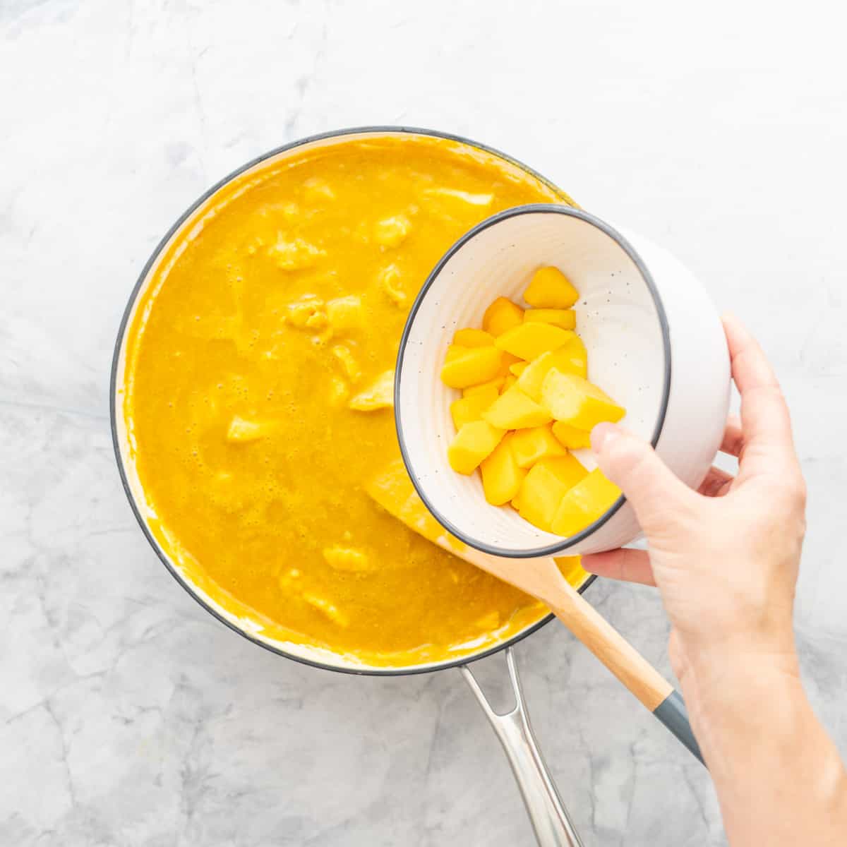 A hand holding a bowl of mango above the pan full of  curry below. 