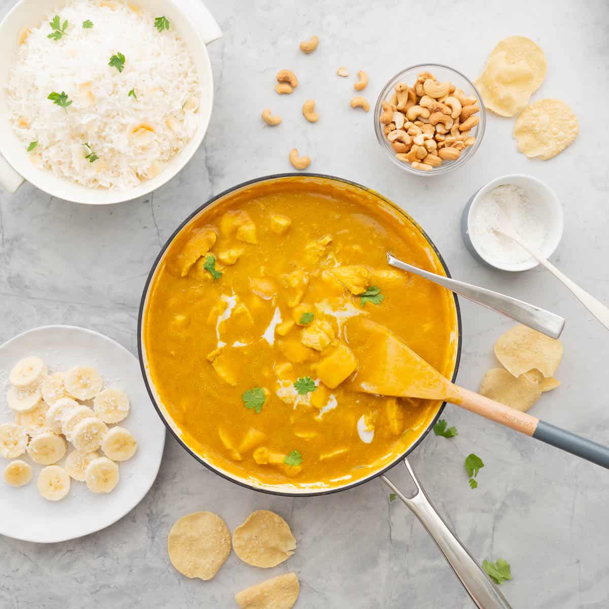 A large pan full of Mango Curry, drizzled with cream and sprinkled with parsley sitting on a bench next to a small bowl of cashew nuts, a ramekin of coconut, a bowl of rice and a plate of sliced bananas and poppadom's scattered over the bench
