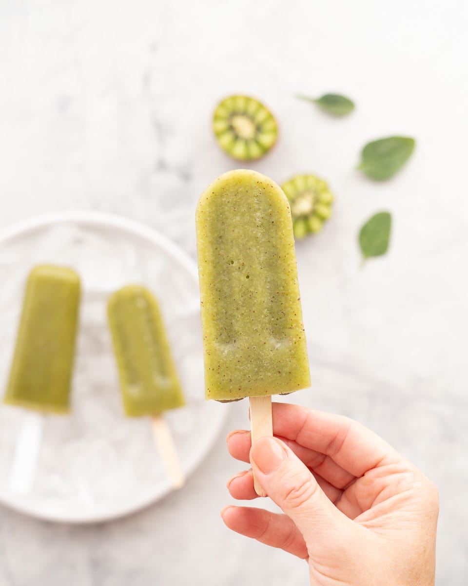 A kiwifruit popsicle held by a hand above a plate of popsicles next to a halved kiwifruit and a scattering of spinach leaves. 