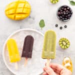 A green kiwi popsicle being held above a plate of brightly coloured fresh fruit and popsicles.
