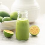 A small glass bottle of green smoothie with a green and white striped straw, a bowl of feijoas in the background.