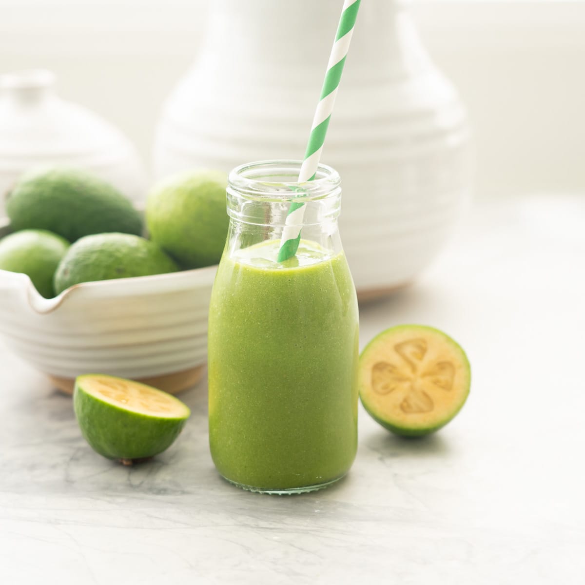 A small glass bottle of green smoothie with a green and white striped straw, a bowl of feijoas in the background. 