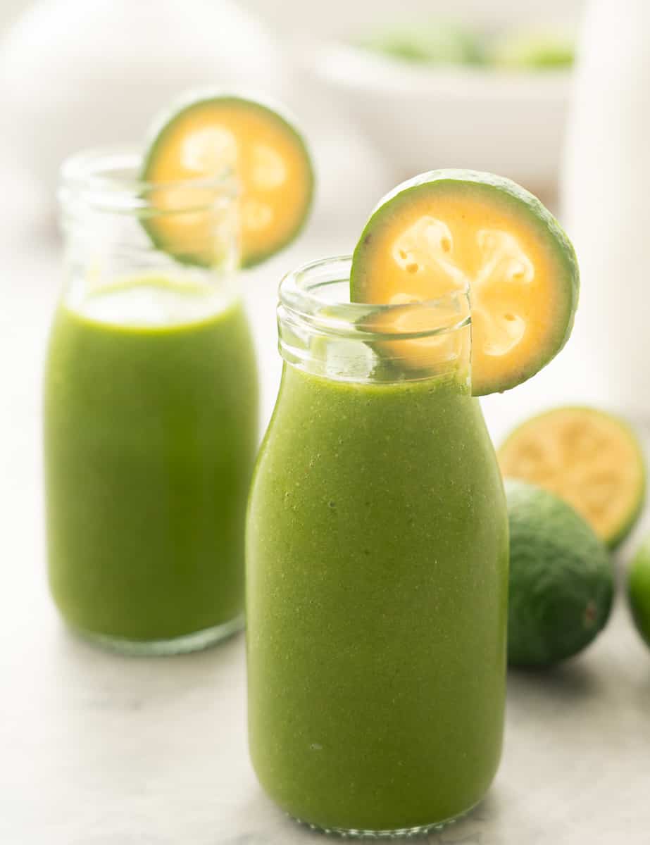 Two small glass bottles filled with green smoothie, the rim of each bottle is garnished with a slice of feijoa. 