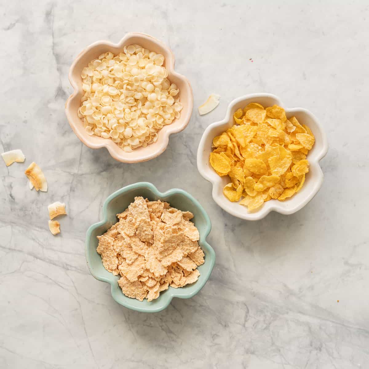 Three small flower shaped ramekins of rice flakes, corn flakes and special k cereal on a marble bench top. 