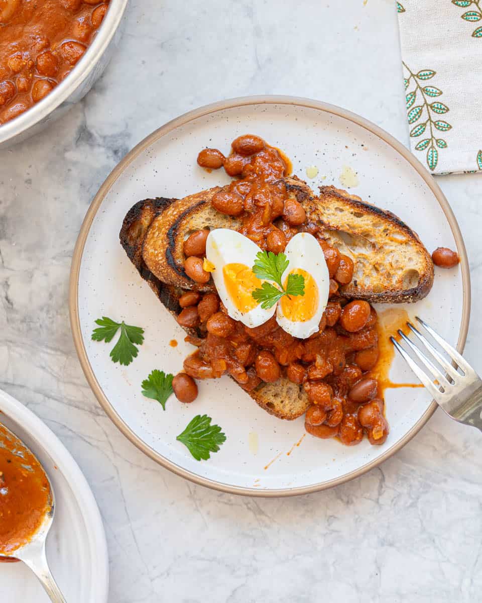 Two slices of rustic toast topped with beans in a rich red sauce and half a hard boiled egg, parsley as garnish. 