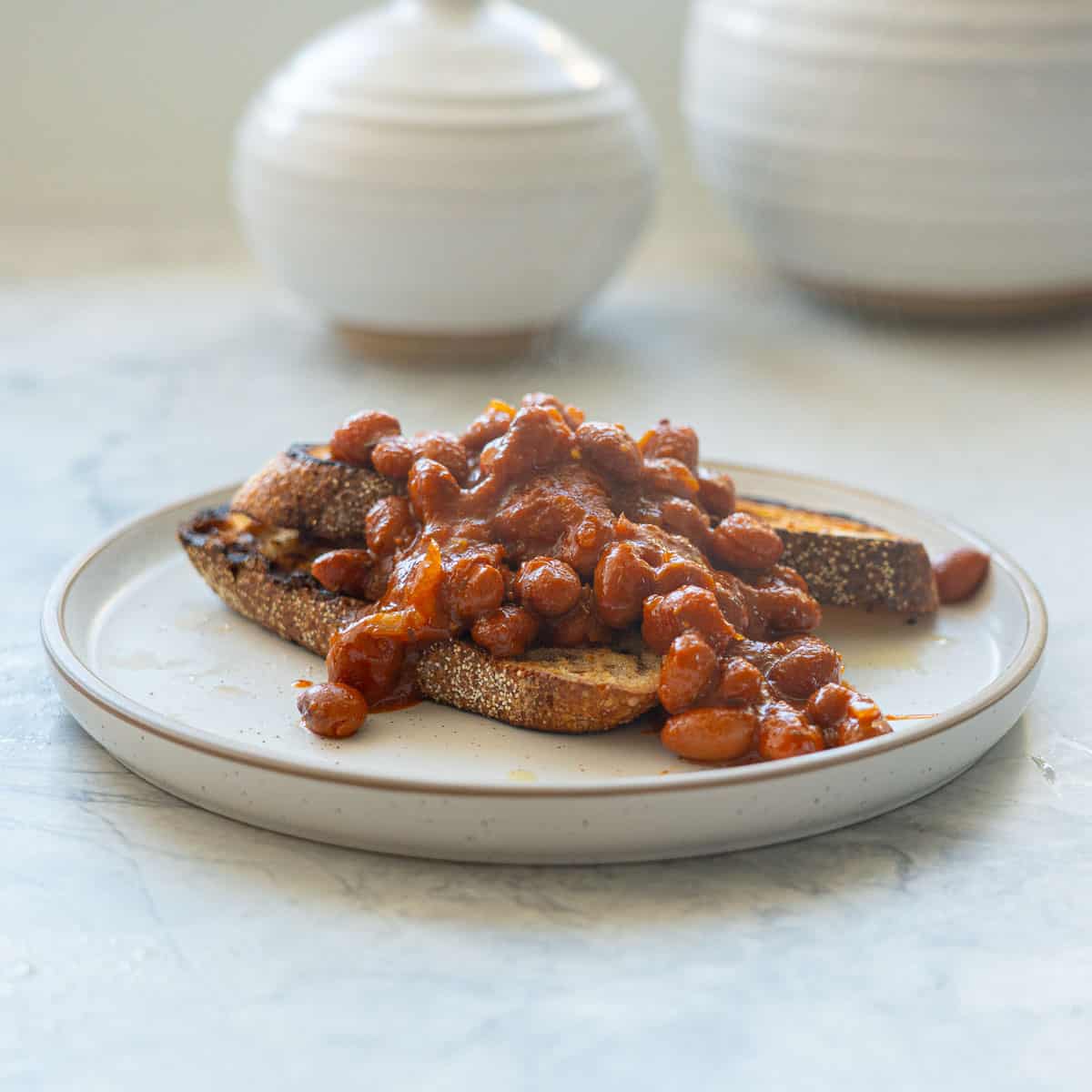 Two slices of rustic toast topped with baked beans on a  white plate with brown rim.