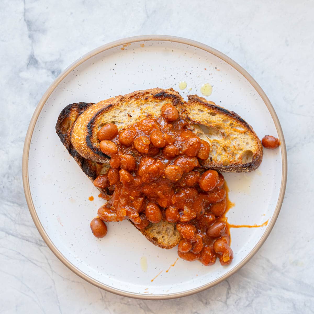 Two slices of rustic toast topped with baked beans on a white plate with brown rim.