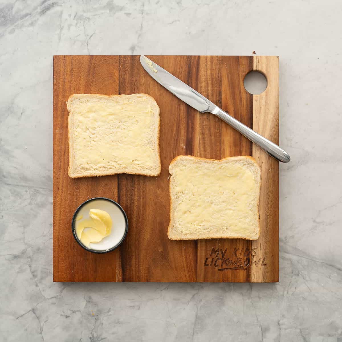 Two slices of buttered bread resting on a wooden chopping board next to a butter knife and a small ramekin of butter. 