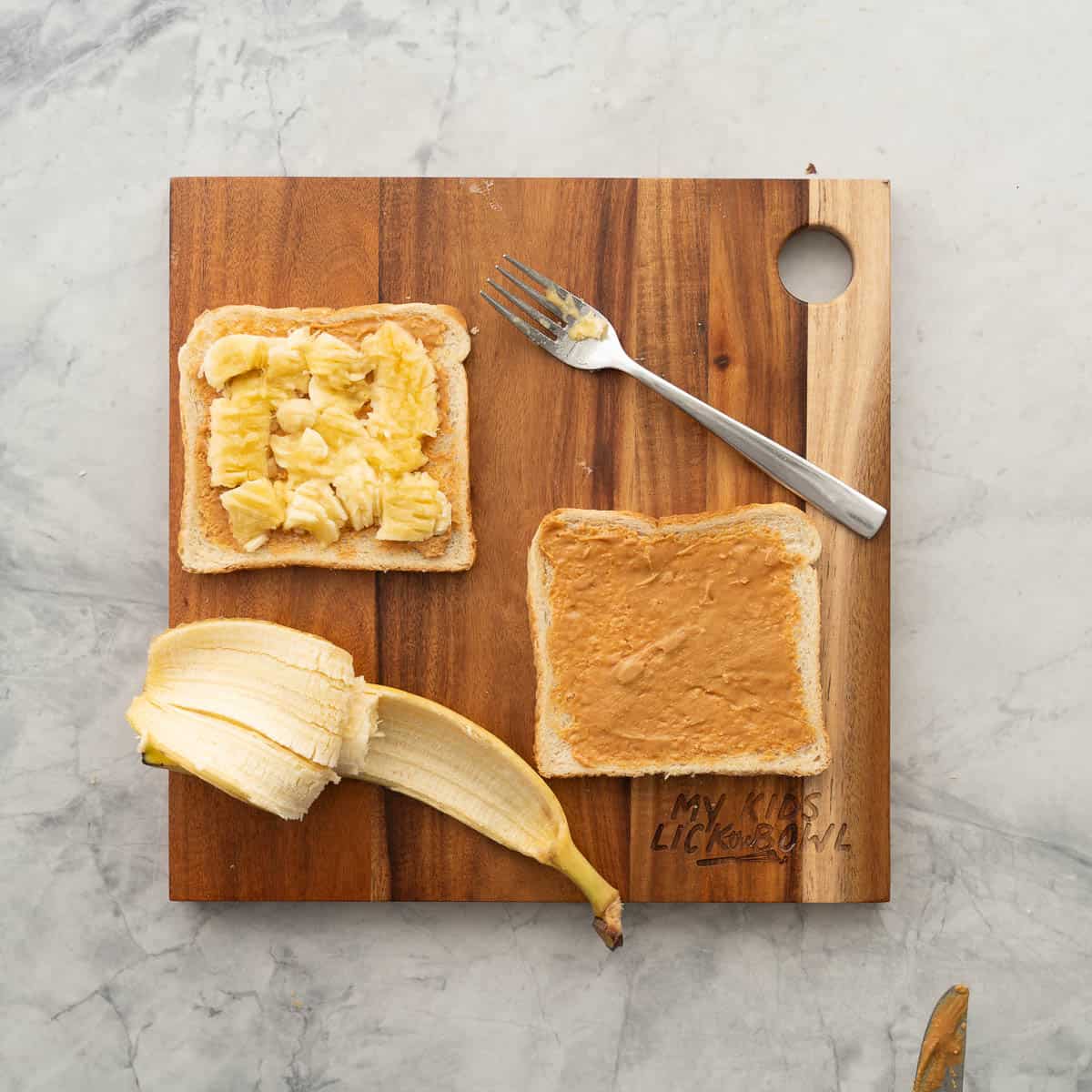Two slices of bread resting on a chopping board and spread with peanut butter. One slice has smashed banana on it. 