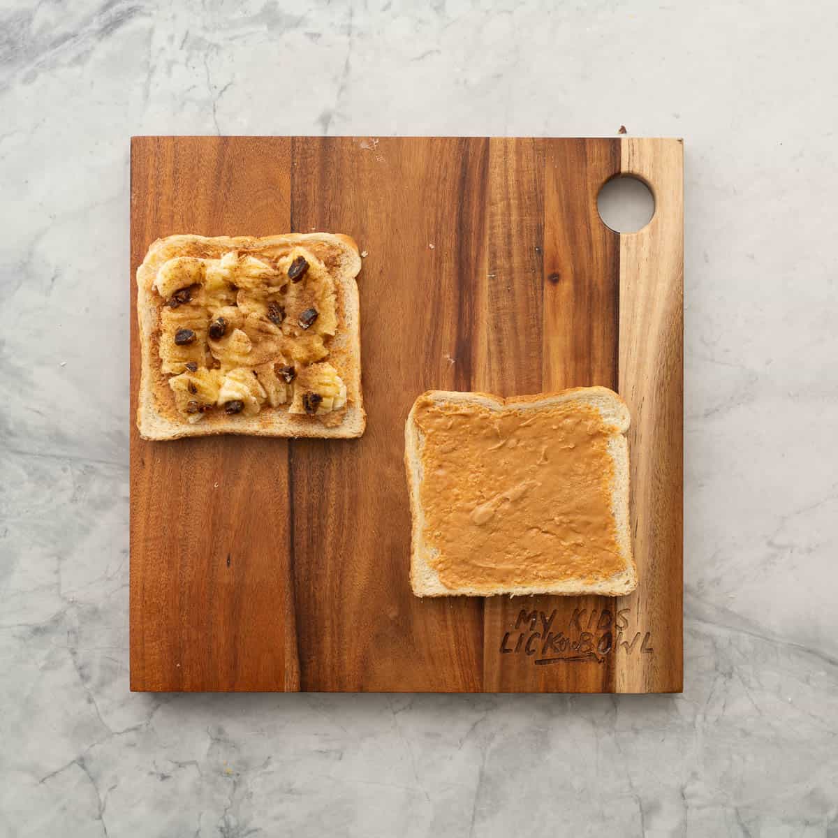 One slice of bread layered with peanut butter, banana and dates and sprinkled with cinnamon resting on a chopping board next to one other slice of bread which is spread with peanut butter. 