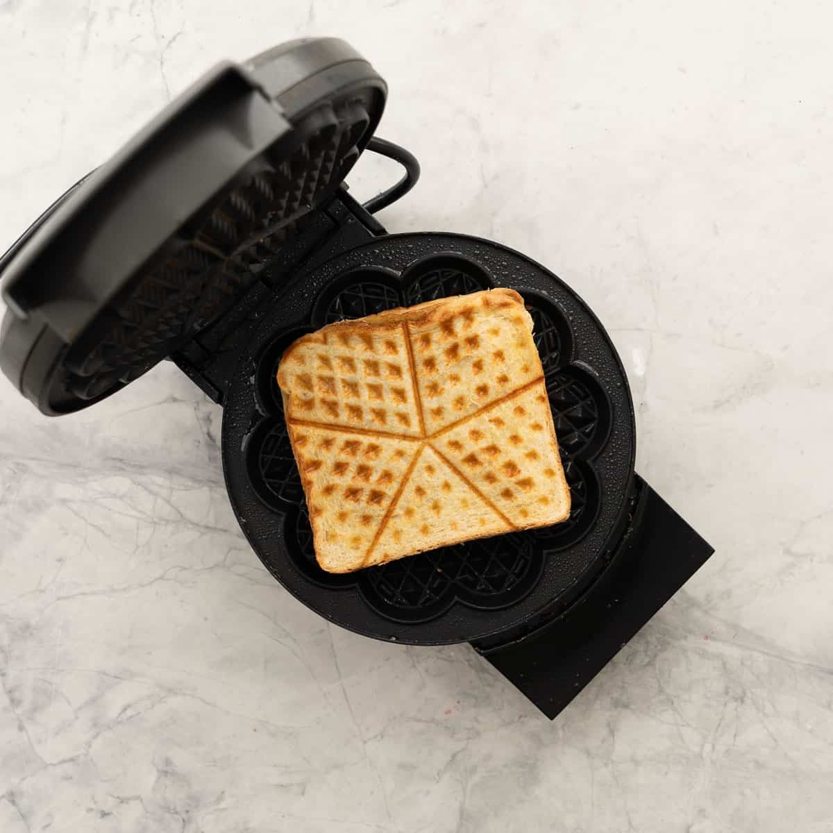 A waffle maker resting on the bench with one toasted peanut butter and banana sandwich