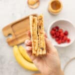 Two grilled peanut butter and banana sandwiches being held up above a chopping board, banana and bowl of raspberries.