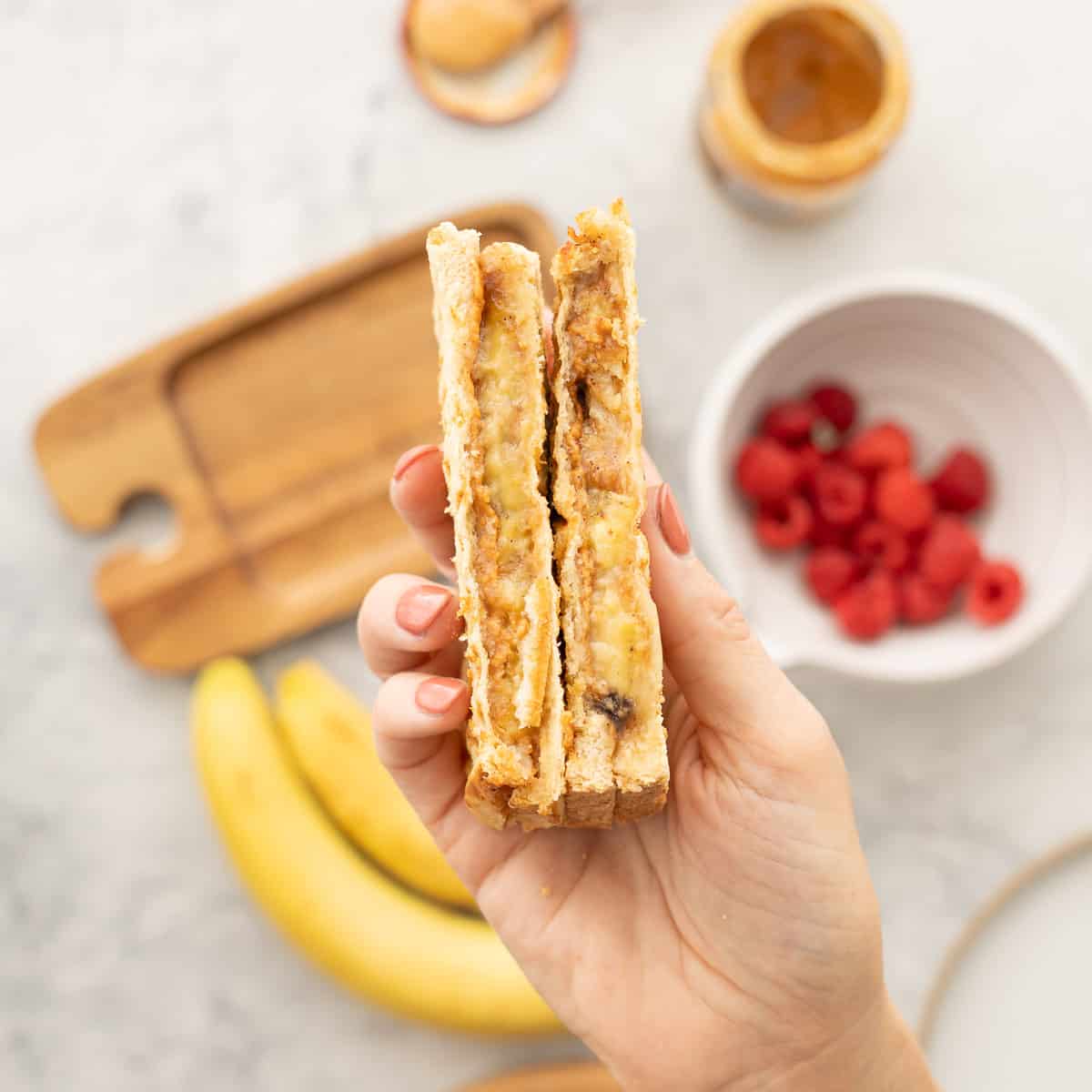 A hand raised above a chopping board holding a halved peanut butter banana sandwich. Next to the wooden chopping board is a ceramic bowl of raspberries and two bananas 