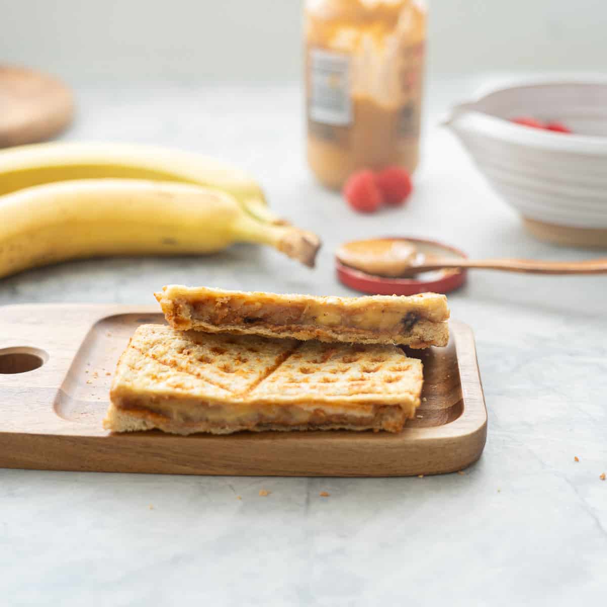 One halved peanut butter banana sandwich resting on a wooden chopping board sitting next to two bananas and a jar of peanut butter 