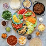 A large selection of taco toppings in and on bright colourful bowls and plates, cut vegetables, beans, mince, tortillas and condiments.