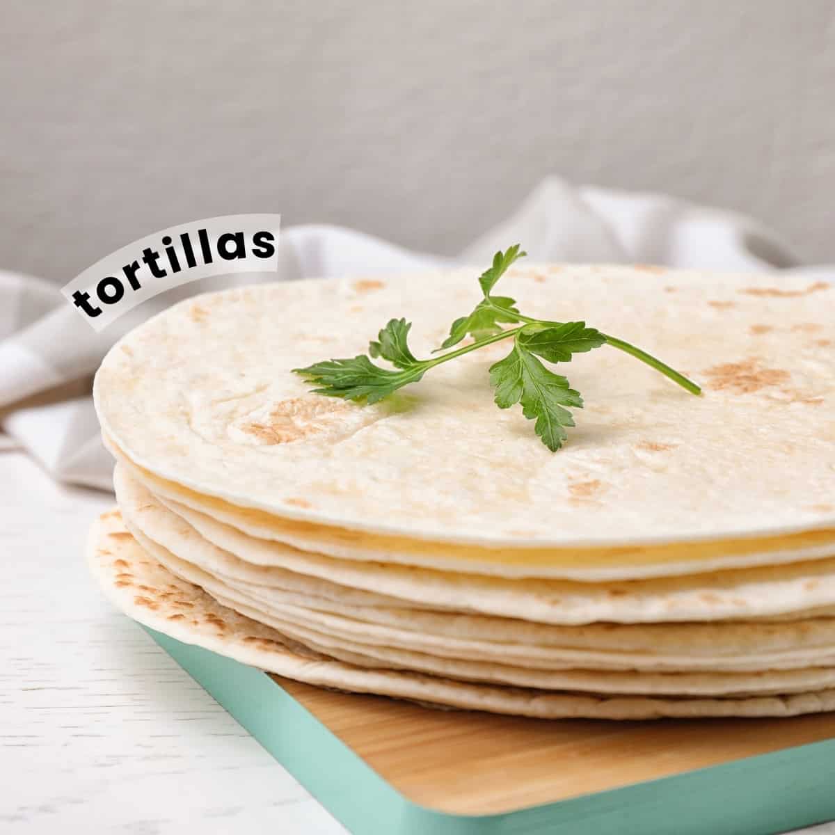 A stack of flour tortillas topped with a garnish of coriander on a wooden chopping board.