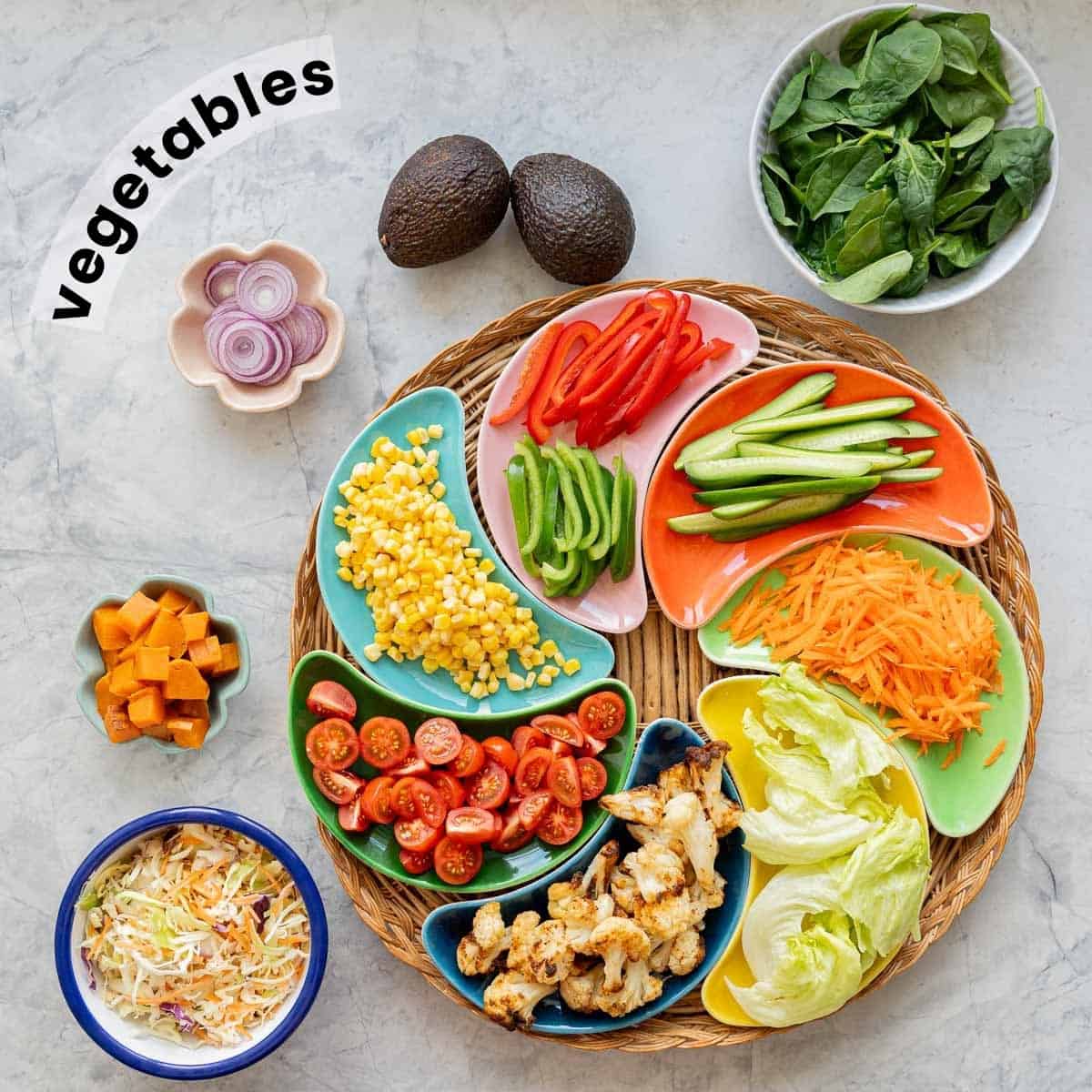 Colourful crescent shaped bowls filled with cut vegetables arranged in a circle on a round wicker tray, with  separate bowls of onion slices, spinach leaves, coleslaw and cubed sweet potato on a bench top. 