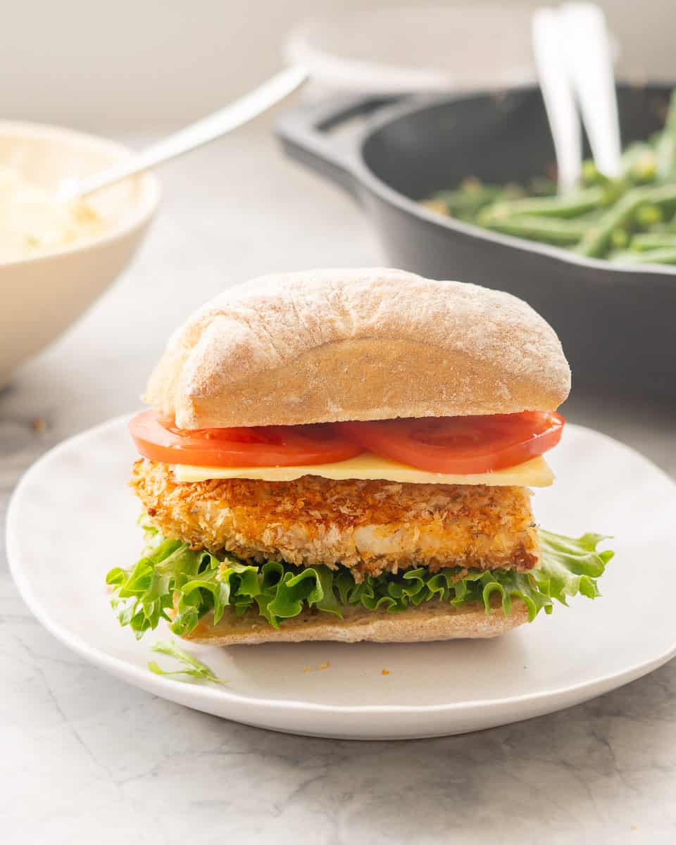 A chicken Birger with tomato slices, cheese, crumbed chicken and lettuce on a small dinner plate. 