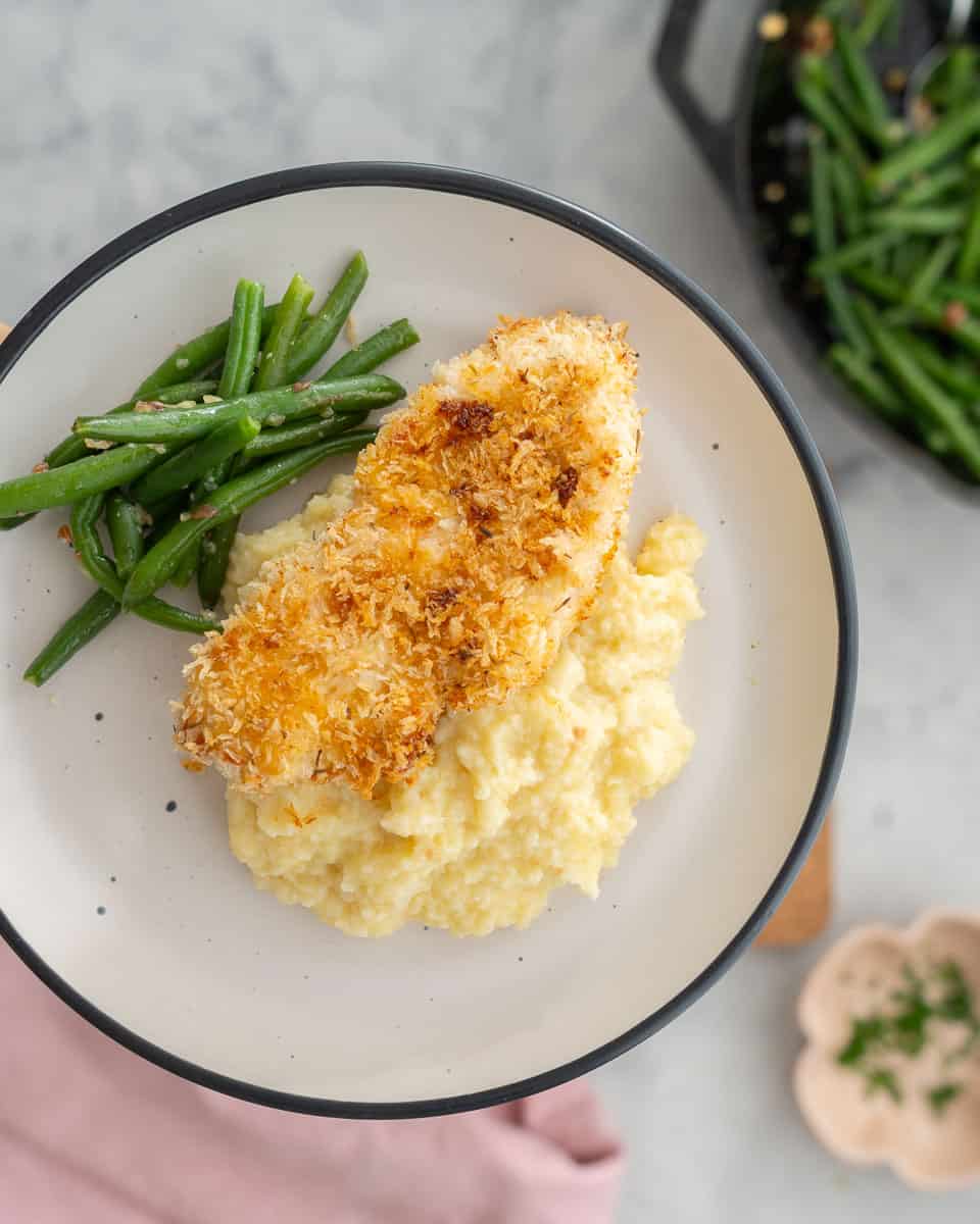 A baked chicken cutlet resting on a bed of mash next to a pile of green beans on a plate. 