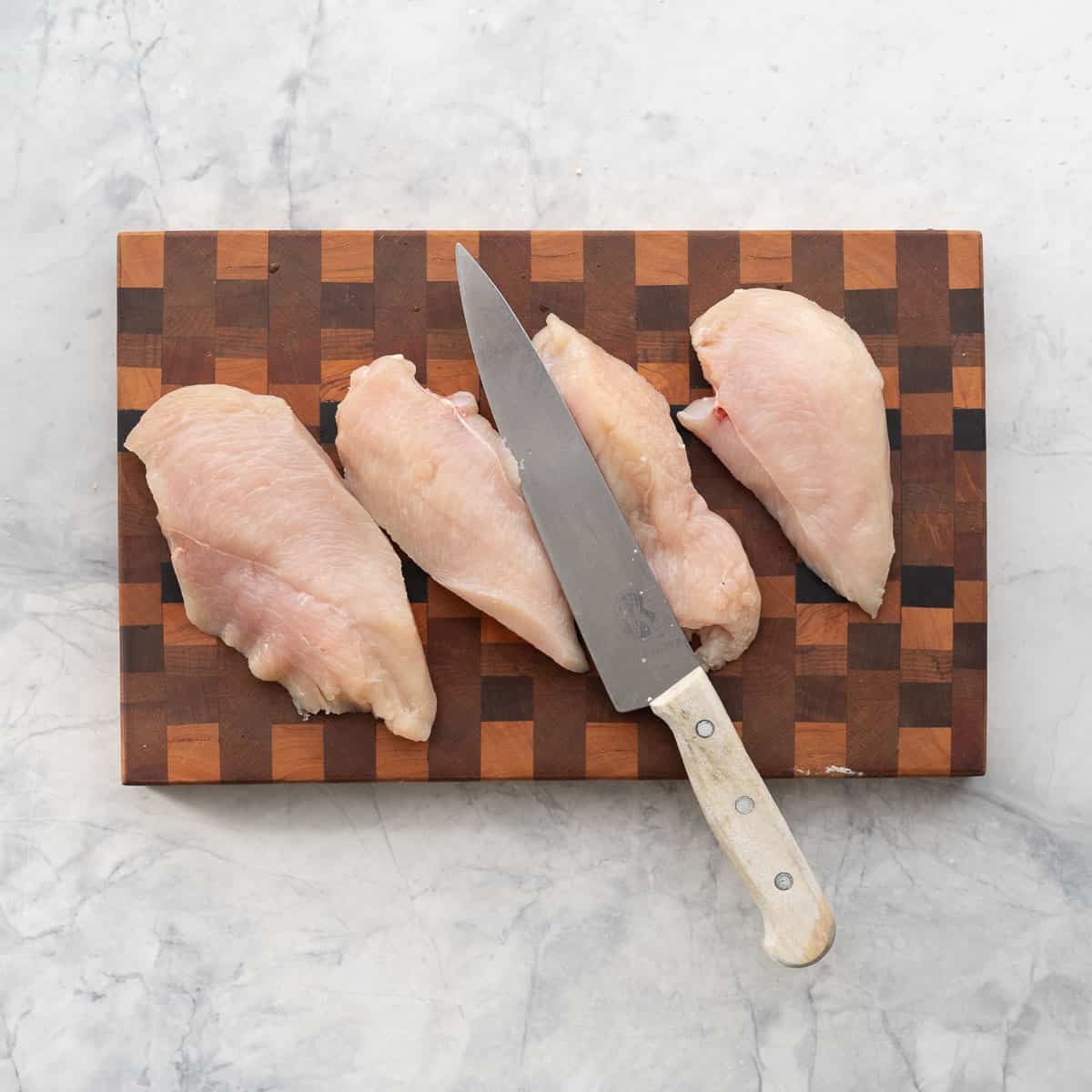 Four chicken cutlets (halved chicken breasts) on a wooden chopping board next to a wooden handled chefs knife. 