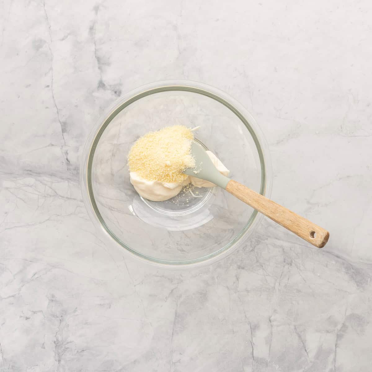 A small glass mixing bowl of mayonnaise and parmesan ready to be mixed together with a small spatula. 