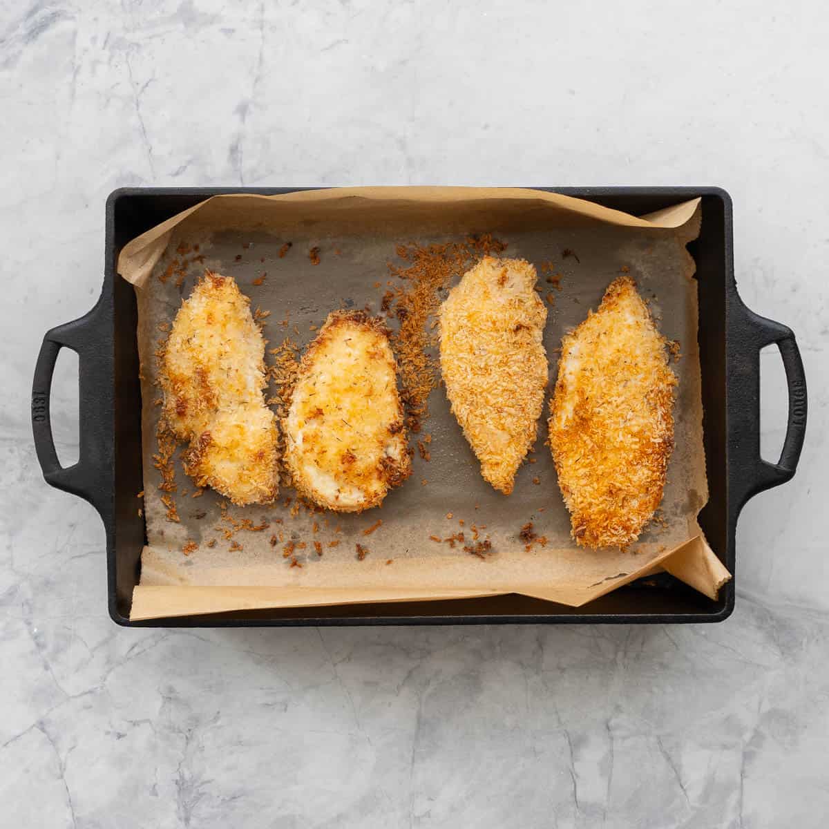 Four cooked chicken cutlets in a rectangular cast-iron baking dish that is lined with brown parchment paper.  